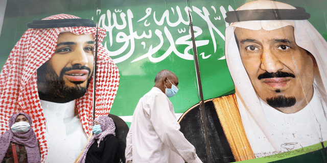 Ludzie noszą maski na twarz, aby chronić przed rozprzestrzenianiem się koronawirusa przed transparentem przedstawiającym króla Arabii Saudyjskiej Salmana, po prawej, i księcia Mohammeda bin Salmana, przed centrum handlowym w Dżuddzie w Arabii Saudyjskiej, w piątek, 5 lutego. 